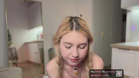 Video of a blonde woman with braided hair, wearing a necklace, looking down with a soft smile in a modern, minimalist kitchen.