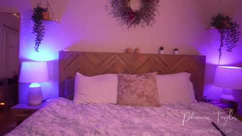 Video of a cozy, dimly-lit bedroom with a wooden headboard, two white pillows, and a floral-patterned throw. Purple mood lighting enhances the rustic decor.