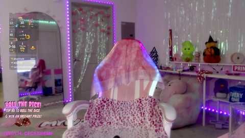 Video of a young woman with pink hair sitting on a pink floral-patterned blanket in a pink chair, surrounded by a bedroom with white walls, a mirror, and stuffed toys.