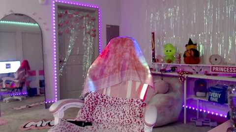 Video of a woman in a pink, leopard-print dress sitting on a white chair, facing a white wall with \"Princess\" and \"Ivy\" signs, surrounded by colorful string lights and decor.