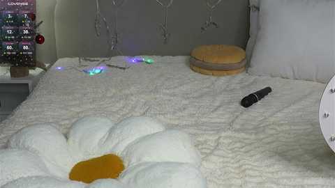 Video of a white bed with a fluffy, yellow daisy pillow, a remote control, and string lights on a gray headboard, creating a cozy, whimsical bedroom scene.