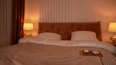 Video of a cozy bedroom with a plush, tufted brown headboard, two white pillows, beige bedding, two lit bedside lamps, and a wooden tray with a remote and glasses on a beige rug.