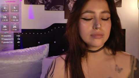 Video of a Latina woman with long, dark hair, wearing a black choker and a nose ring, lying on a bed with a black headboard. Background includes monochrome posters and purple lighting.