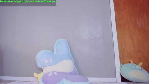 A video showing a plush toy with a blue and purple body, resembling a dolphin, lying on a wooden floor. Text in the top left corner reads, \"Please do not forget to leave a thumbs up, thank you.\
