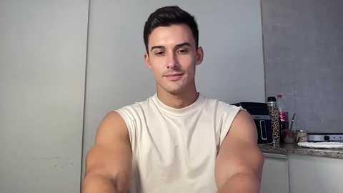 Video of a muscular, light-skinned man with short dark hair, wearing a sleeveless white shirt, in a modern kitchen with a granite countertop, utensils, and a toaster.