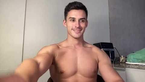 Video of a shirtless, muscular young man with short dark hair, smiling, in a modern kitchen with a granite countertop and appliances, including a microwave and green towels.