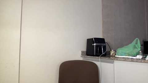 Video of a minimalist kitchen corner with a beige wall, a black toaster, a green cloth, and a brown chair.
