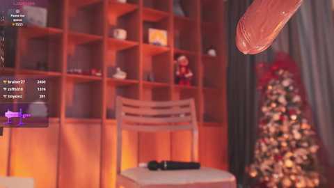 Video of a cozy, dimly lit room with wooden shelves, a Christmas tree, and a man with a large, erect penis in front of a video game screen.