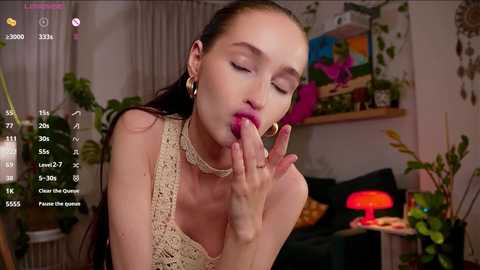 A video of a fair-skinned, slender woman with long brown hair, wearing a beige crochet top, licking her fingers in a cozy, plant-filled room with colorful artwork and a red lamp.