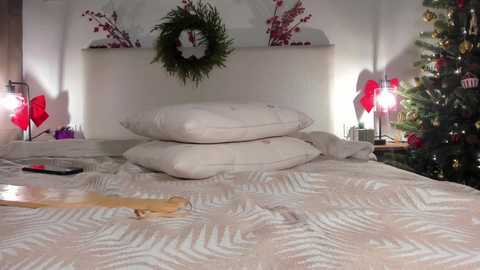 Video of a cozy, decorated bedroom with a white headboard, Christmas tree, wreath, and a white geometric-patterned bedspread.