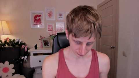 Video of a young man with short brown hair, wearing a red tank top, standing in a cozy, softly lit room with beige walls, black office chair, floral decorations, and anatomical posters.