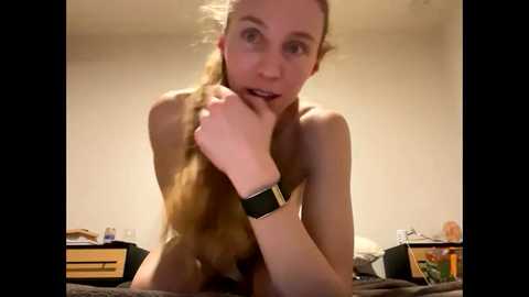 Media: Video of a young, fair-skinned, slender woman with long blonde hair, topless, wearing a black smartwatch. She's in a dimly lit bedroom with beige walls, messy desk, and a bed.