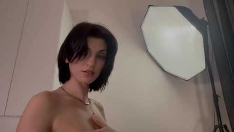 A video of a fair-skinned woman with short black hair, topless, covering her small breasts with one hand. She wears a gold necklace. Background includes a hexagonal light reflector and a dark curtain.