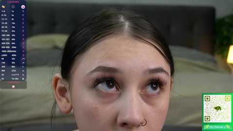 Video of a young woman with fair skin, brown eyes, and dark hair pulled back, wearing a septum ring. Background shows a grey bed and green plant.