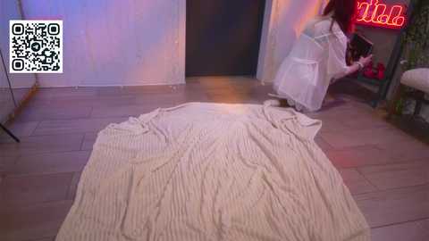 Video of a dimly lit room with a white knit blanket on a wooden floor. A person in a white dress with a black belt is bent over, seemingly picking up something. QR code in the top left corner.