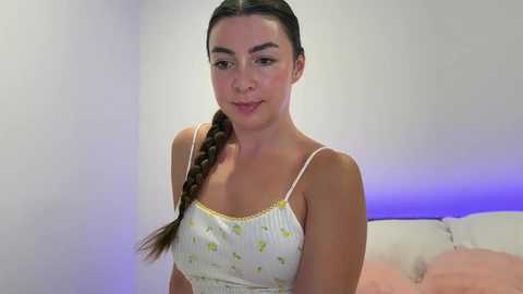 Media: Video of a young woman with fair skin and dark hair in a braid, wearing a white camisole with yellow flowers, sitting on a beige sofa with a pink blanket.