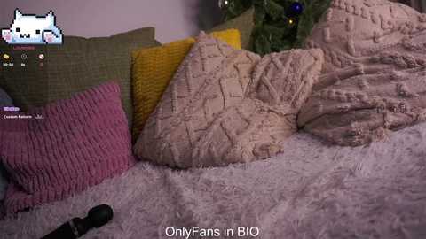 Video of a cozy, plush bed with a white furry blanket, yellow and pink knitted pillows, and a small Christmas tree in the background. Text reads \"OnlyFans in BIO.\