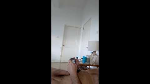 A video of a person holding a phone, wearing light-colored clothes, lying on a bed in a bright, minimalist room with white walls and doors.