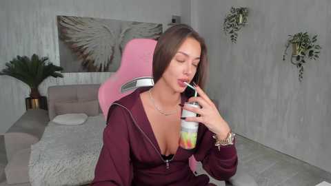 Media: Video of a woman with long brown hair, wearing a low-cut maroon hoodie, drinking from a colorful straw, in a modern, minimalist bedroom with gray walls and greenery.