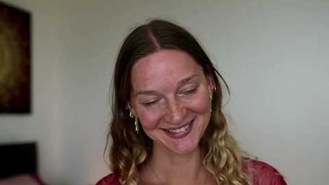 A video of a smiling woman with light brown hair in braids, wearing a red top, standing against a plain white wall with a framed picture in the background.
