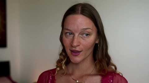 Video of a fair-skinned, brunette woman with wavy hair, wearing a red blouse, standing against a beige wall with a blurred dark painting in the background.