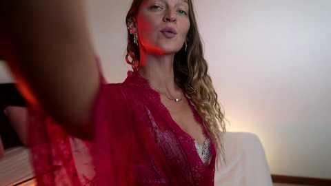 Video of a woman with long, wavy blonde hair, wearing a red lace dress, looking pensive, against a blurred background with red lighting.