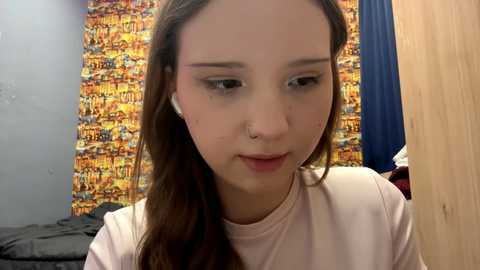 Video of a young woman with long brown hair, wearing headphones and a white top, looking down, against a colorful, abstract wall background.