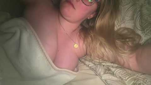 A video of a young Caucasian woman with long blonde hair, wearing glasses and a yellow necklace, lying on a beige, quilted bedspread. She is partially covered by a white blanket.