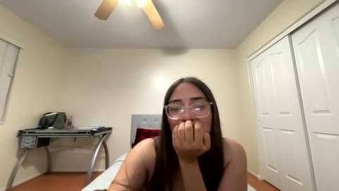 Video of a woman with long brown hair and glasses, sitting at a desk, covering her mouth, in a beige-walled room with a white door and ceiling fan.
