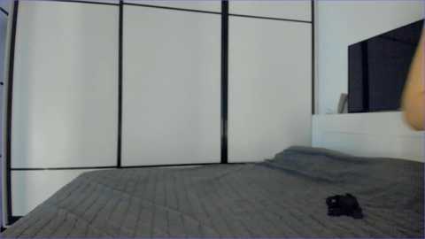 A video of a minimalist bedroom with a bed covered in a grey quilted blanket, a black garment tossed on the bed, and a sliding panel partition with black frames separating the space.