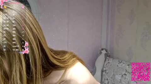 Video of a young woman with long, straight, light brown hair, seen from behind. She is wearing a light-colored tank top. The background features a floral-patterned wallpaper and a white bed with a white pillow. The image includes a pink overlay with a watermark reading \"LIVE STREAM.\