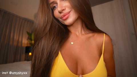 Video of a young woman with long, straight brown hair, wearing a low-cut yellow spaghetti-strap top, standing in a dimly-lit bedroom. She has a light skin tone and a subtle smile.