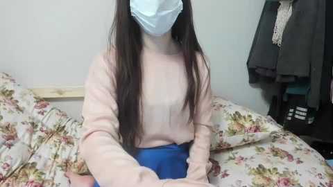 Video of a young woman with long brown hair, wearing a light pink sweater, blue skirt, and a blue surgical mask, sitting on a bed with a floral bedspread, in a simple, cluttered bedroom.