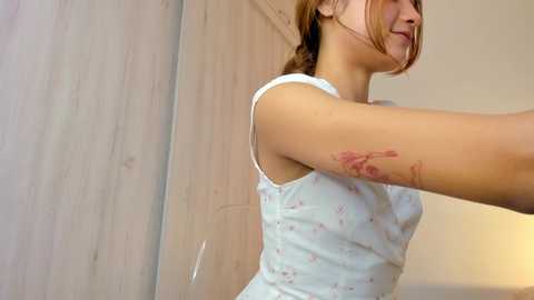 A video of a fair-skinned woman with light brown hair, wearing a light blue tank top, with a red bird tattoo on her upper arm. She's in a softly lit room with light-colored walls and a wooden door.