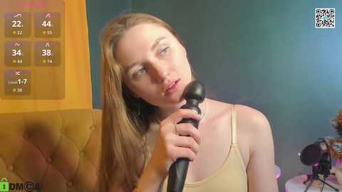 A video of a young, fair-skinned woman with long blonde hair, wearing a beige tank top, holding a black microphone while singing. Background includes a green wall and a colorful digital clock.