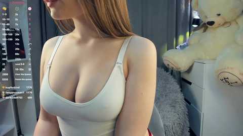 A video of a woman with fair skin and long, straight, light brown hair, wearing a tight, white tank top that accentuates her large breasts. She stands in a room with a gray curtain, a white dresser, and a plush teddy bear.