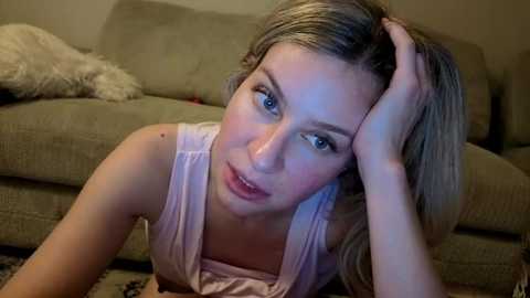 Video of a young woman with light skin, blue eyes, and shoulder-length blonde hair, lying on a beige carpet in a living room. She wears a white tank top and appears tired, resting her head on her hand.