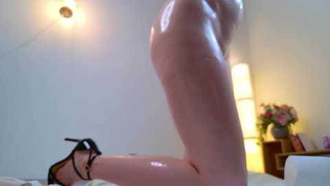 Media: Video of a nude woman kneeling on a bed, showcasing her fair skin, slender physique, and wearing black high heels. Background features a white wall, a tall floor lamp, and a bouquet of flowers on a wooden nightstand.