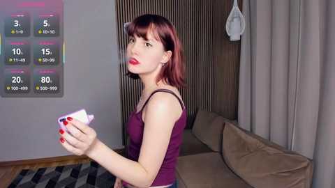 Media: Video of a red-haired woman in a purple top holding a white card, standing in a modern living room with a beige couch, light wooden walls, and a digital temperature display on the left.