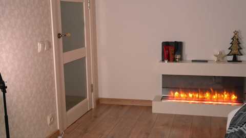Video of a minimalist bedroom with a white door, beige wallpaper, wooden floor, modern white fireplace with glowing embers, bookshelf with books and Christmas ornaments, and a partial view of a bed.