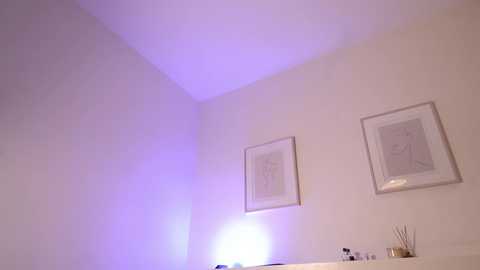 Video of a minimalist, white-walled bathroom with two framed minimalist drawings, a glowing blue light, and a white sink with a few items.