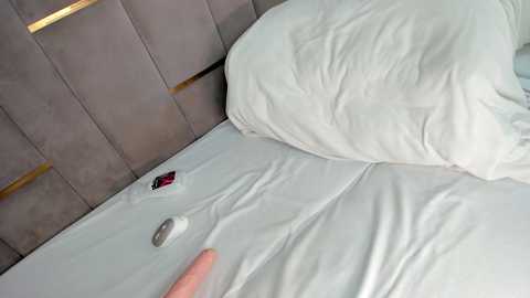Video of a person lying in bed with white sheets, a gray wall with gold trim in the background, a red alarm clock on the bedside table, and a hand reaching out.