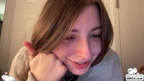 A video of a young Caucasian woman with light brown hair, smiling while playfully pulling her hair. She wears a light grey sweater. The background shows a white door.