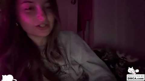 A dimly-lit video of a young woman with long brown hair, wearing a graphic T-shirt, in a dimly-lit room with a purple light.