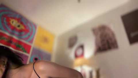 Video of a woman with medium skin tone, partially visible, in a colorful, psychedelic room with wall art, a lamp, and a blurred background.