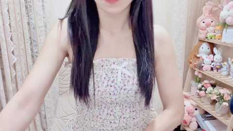 Video of an Asian woman with long black hair, wearing a strapless, floral-patterned dress. She stands in a pastel-colored room with a beige quilted wall, pink bunny plushies, and a wooden shelf filled with more plush toys.