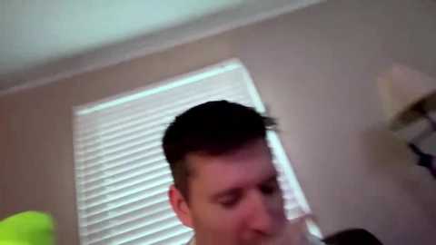 A video of a man with short brown hair, wearing a black shirt, looking down. The background features a white window blind and a beige wall.