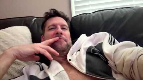 Video of a middle-aged man with short dark hair and a beard, lying on a black leather couch. He is shirtless, wearing a white and black striped polo, with a hand near his mouth. The background shows a beige wall and white blinds.