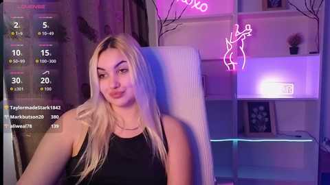 Video of a young woman with long blonde hair, wearing a black tank top, seated in a modern living room with neon lights, shelves, and a framed picture.