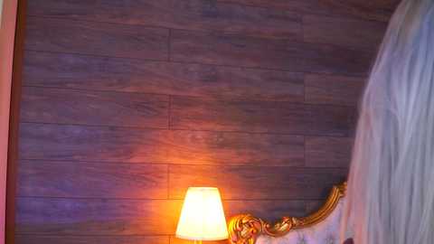 Video of a bed with a wooden headboard, featuring a golden, ornate design. A lamp with a white shade emits a warm glow, casting a soft light on the dark brown, textured wooden wall. The background includes a sheer white curtain hanging on the right.